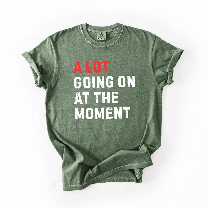 May include: Olive green t-shirt with the text "A LOT GOING ON AT THE MOMENT" in white and red lettering. The shirt is folded and has a crew neck.