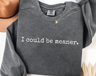 May include: Dark gray sweatshirt with the phrase "I could be meaner" in white lettering. A gold necklace with a pendant is draped on the shirt. A brown hat is visible in the background.
