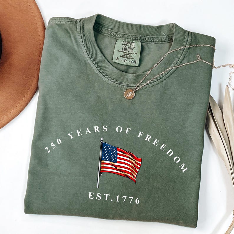 Pu&ograve; includere: T-shirt verde oliva con la scritta "250 YEARS OF FREEDOM" e una grafica della bandiera americana. La maglietta ha anche la scritta "EST. 1776". Una collana d'oro con un ciondolo &egrave; sulla maglietta. Un cappello marrone &egrave; sullo sfondo.