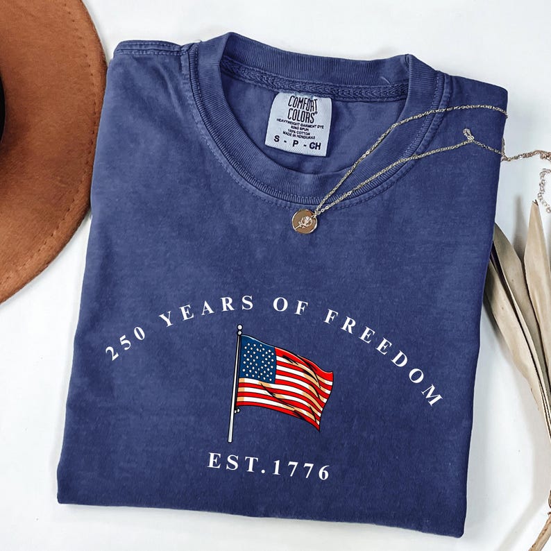 Pu&ograve; includere: Una maglietta blu navy con la scritta "250 YEARS OF FREEDOM" e una grafica della bandiera americana. La maglietta presenta anche la scritta "EST. 1776". Una collana d'oro con un ciondolo &egrave; drappeggiata sulla maglietta.
