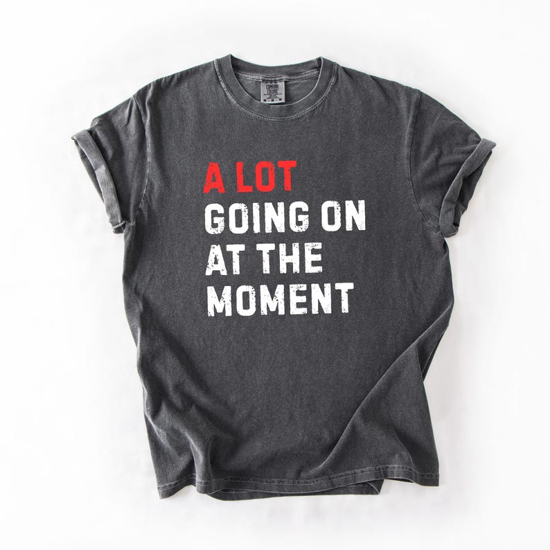 May include: Dark gray t-shirt with the phrase "A LOT GOING ON AT THE MOMENT" printed in white and red. The shirt has a crew neck and short sleeves, with a slightly faded, vintage look.