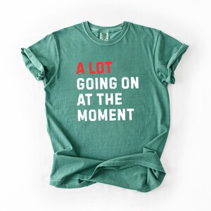 May include: A green t-shirt with the text "A LOT GOING ON AT THE MOMENT" in white and red block letters. The shirt is folded and has rolled-up sleeves. The shirt is made of a soft material.