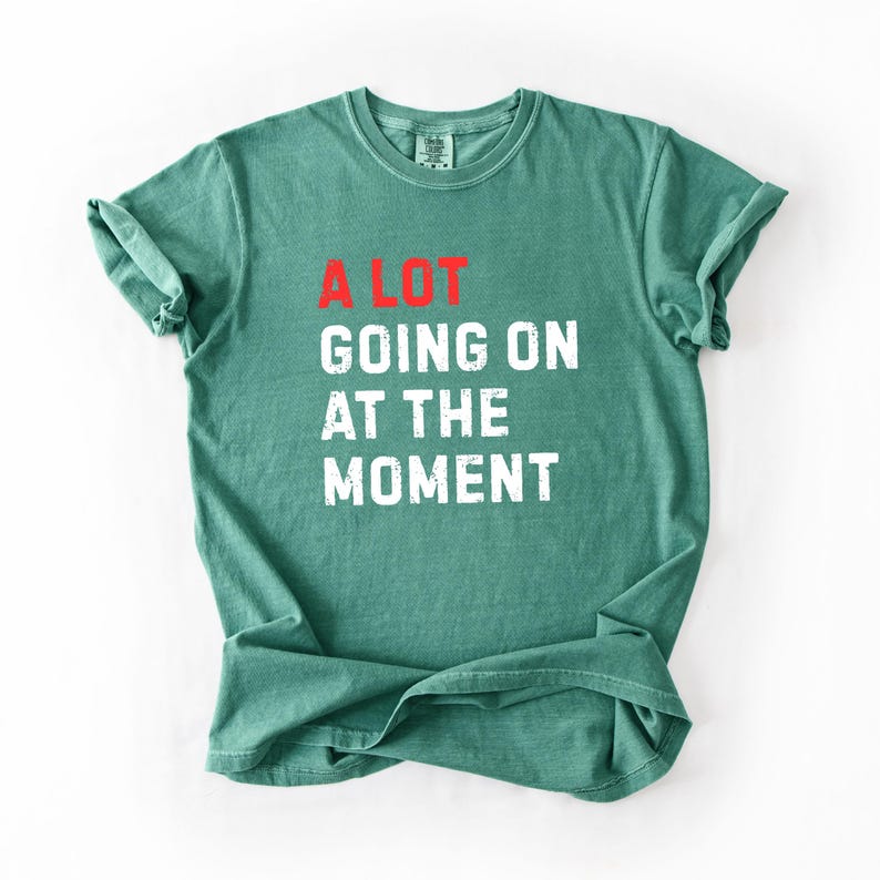 May include: A green t-shirt with the text "A LOT GOING ON AT THE MOMENT" in white and red block letters. The shirt is folded and has rolled-up sleeves. The shirt is made of a soft material.