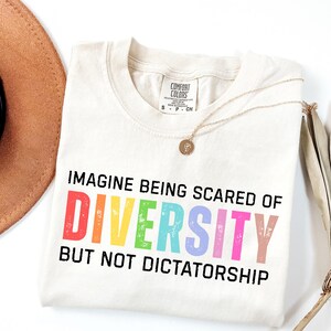 May include: Cream-colored t-shirt with the text "IMAGINE BEING SCARED OF DIVERSITY BUT NOT DICTATORSHIP" in colorful letters. A gold necklace and a brown hat are also visible.