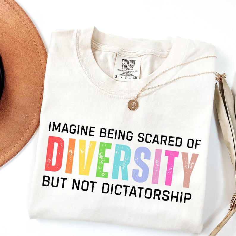 May include: Cream-colored t-shirt with the text "IMAGINE BEING SCARED OF DIVERSITY BUT NOT DICTATORSHIP" in colorful letters. A gold necklace and a brown hat are also visible.