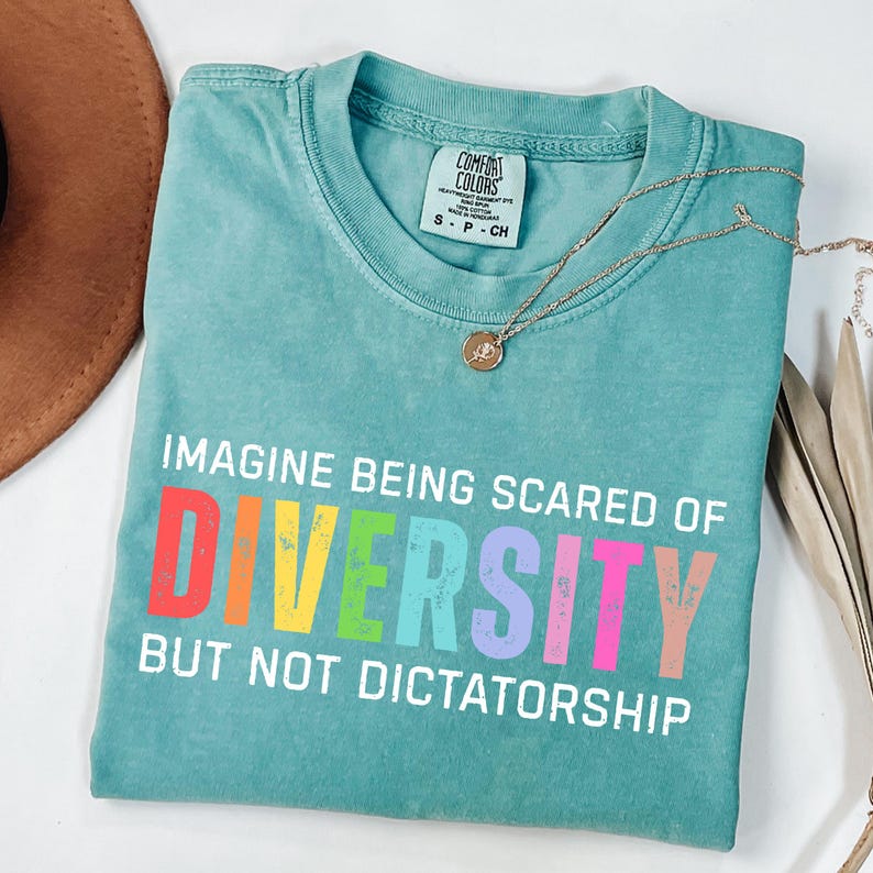 May include: A turquoise Comfort Colors t-shirt with the text "IMAGINE BEING SCARED OF DIVERSITY BUT NOT DICTATORSHIP" in white and rainbow colors. A gold necklace with a pendant is on the shirt. The shirt is folded on a white surface.