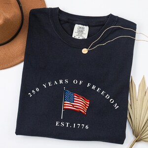 Pu&ograve; includere: T-shirt blu navy con la scritta "250 YEARS OF FREEDOM" e una grafica della bandiera americana. La maglietta presenta anche la scritta "EST. 1776". Sono visibili anche una collana d'oro e un cappello marrone.