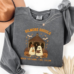 May include: Dark gray sweatshirt with the text "GILMORE GHOULS" in orange. The design features two ghost figures under a gazebo, pumpkins, bats, and the phrase "WHERE YOU HAUNT, I WILL FOLLOW". A brown hat and necklace are visible.