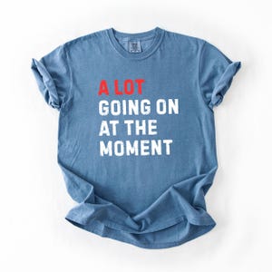 May include: Blue t-shirt with the text "A LOT GOING ON AT THE MOMENT" in white and red block letters. The shirt is made of a soft material and has short sleeves.