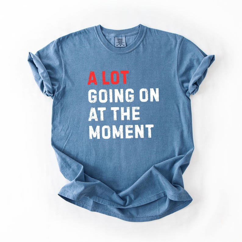 May include: Blue t-shirt with the text "A LOT GOING ON AT THE MOMENT" in white and red block letters. The shirt is made of a soft material and has short sleeves.