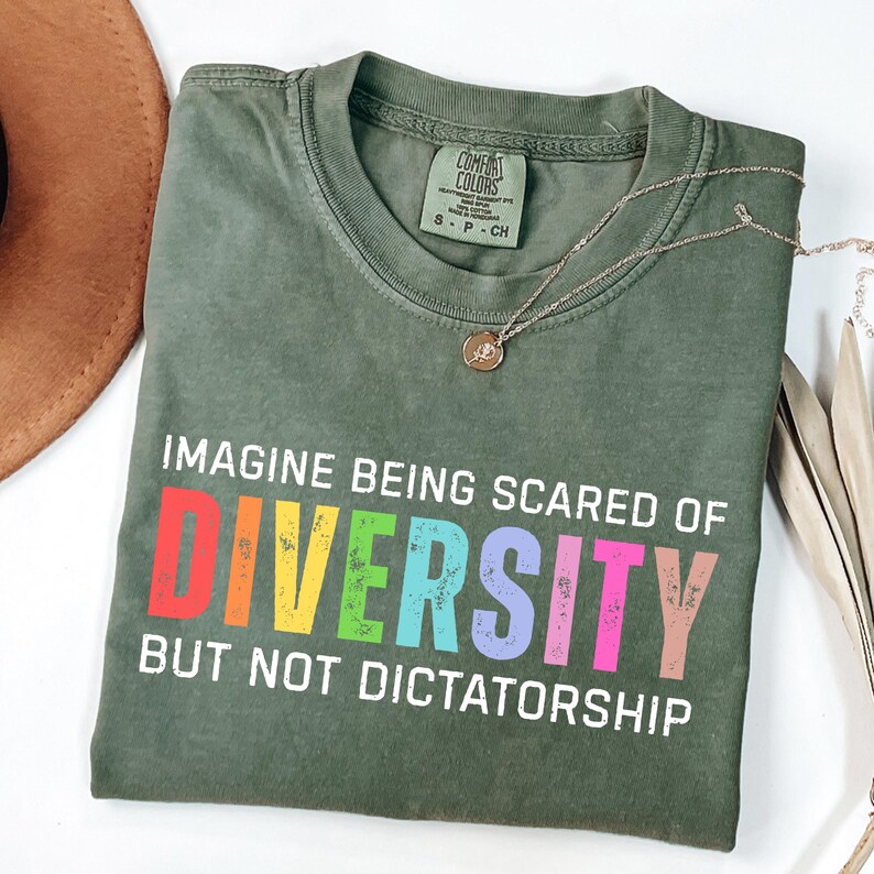 May include: An olive green t-shirt with the text "IMAGINE BEING SCARED OF DIVERSITY BUT NOT DICTATORSHIP" in white and rainbow colors. A gold necklace with a pendant is on the shirt. A brown hat is visible in the upper left corner.