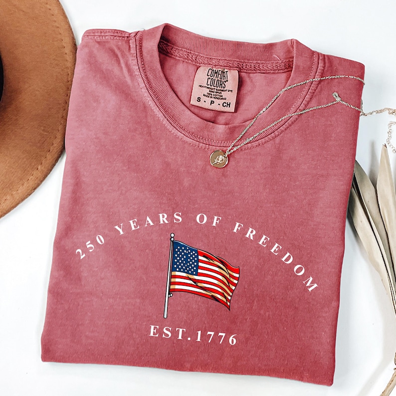Pu&ograve; includere: T-shirt color rosa polvere con la scritta "250 YEARS OF FREEDOM" e un'illustrazione della bandiera americana. La scritta "EST. 1776" &egrave; sotto la bandiera. Una collana d'argento con un piccolo ciondolo &egrave; sulla maglietta.