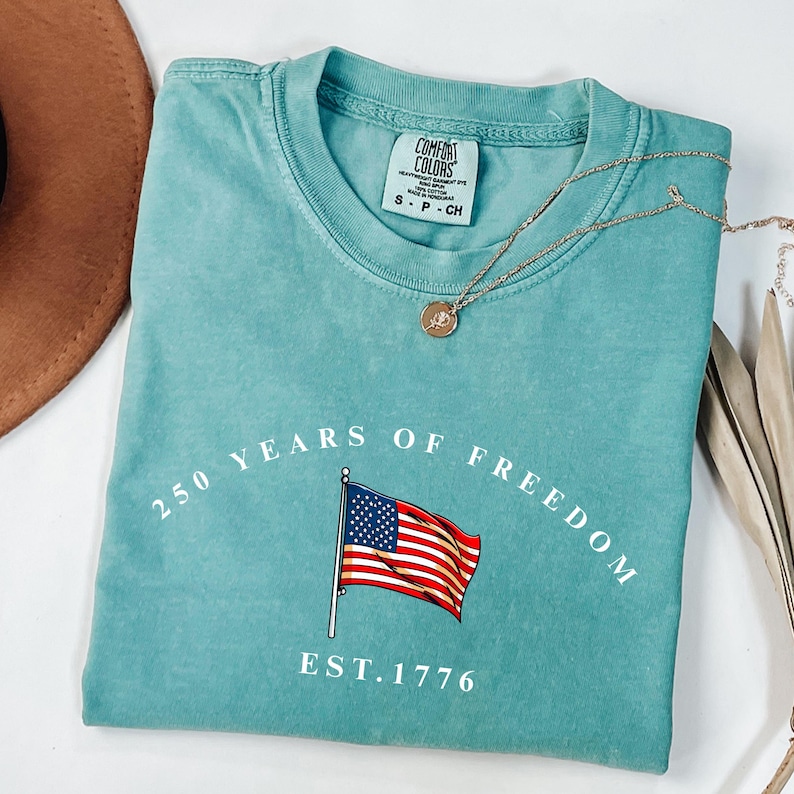 Pu&ograve; includere: Una maglietta turchese con la scritta "250 YEARS OF FREEDOM" e una grafica della bandiera americana. Il testo "EST. 1776" &egrave; sotto la bandiera. Una collana d'oro con un ciondolo &egrave; sulla maglietta.