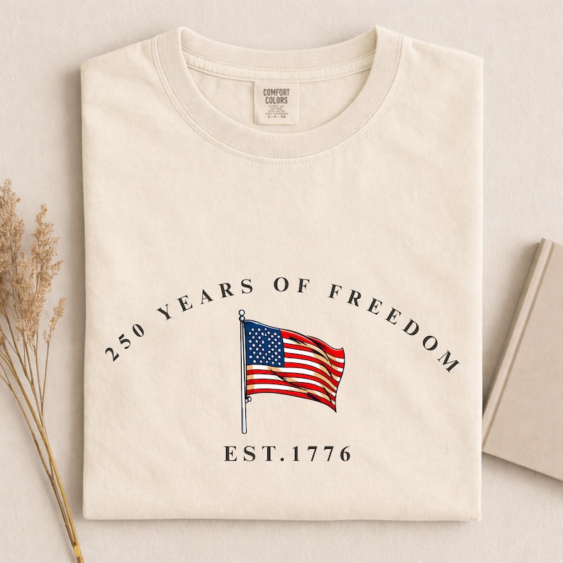 Pu&ograve; includere: T-shirt color crema con grafica della bandiera americana e la scritta "250 YEARS OF FREEDOM EST. 1776". La maglietta &egrave; piegata e ha un'etichetta Comfort Colors.