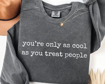 May include: Dark gray sweatshirt with the text "you're only as cool as you treat people" in white. A gold necklace with a pendant is draped on the shirt. A brown hat is visible in the background.