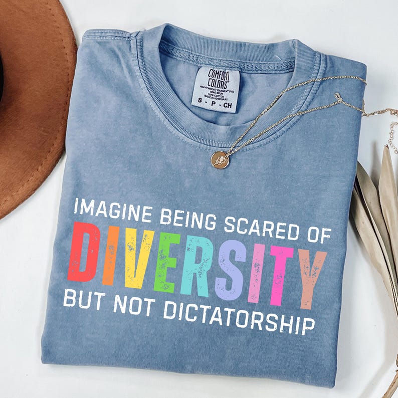 May include: A blue Comfort Colors t-shirt with the text "IMAGINE BEING SCARED OF DIVERSITY BUT NOT DICTATORSHIP" in white and rainbow colors. A gold necklace with a pendant is draped across the shirt.