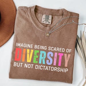 May include: A brown Comfort Colors t-shirt with the text "IMAGINE BEING SCARED OF DIVERSITY BUT NOT DICTATORSHIP" in colorful letters. A silver necklace with a pendant is draped across the shirt.
