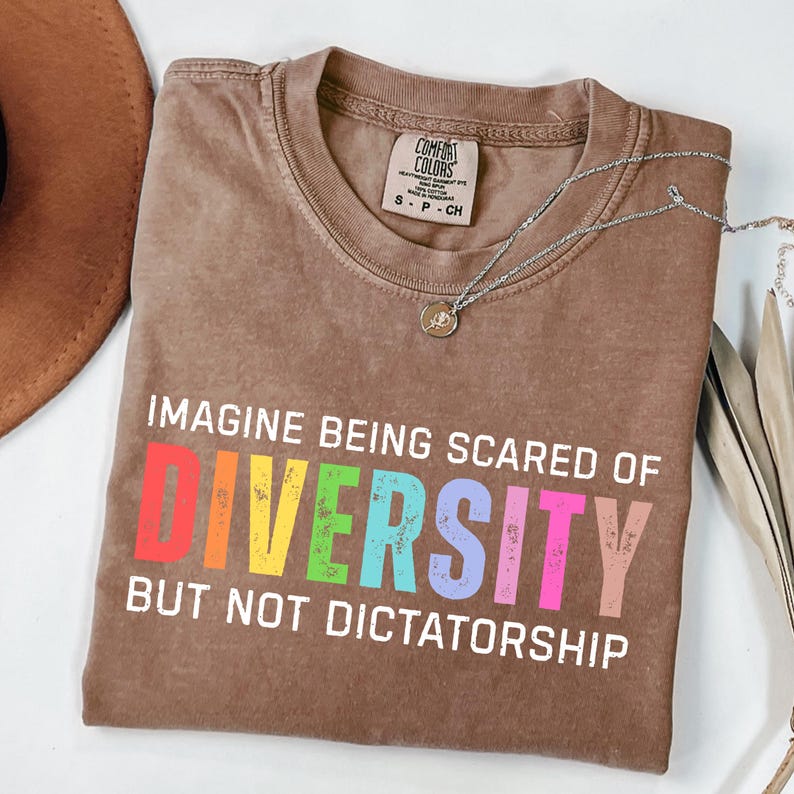 May include: A brown Comfort Colors t-shirt with the text "IMAGINE BEING SCARED OF DIVERSITY BUT NOT DICTATORSHIP" in colorful letters. A silver necklace with a pendant is draped across the shirt.