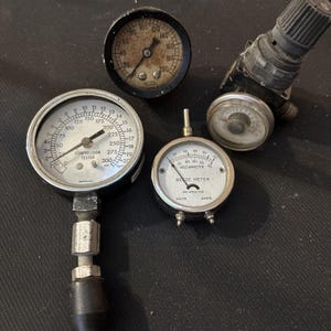 May include: A collection of vintage mechanical gauges including a large compression tester with a 300 psi scale, a round pressure gauge with a 200 psi range, a circular voltammeter labeled "Bede Meter" for volts and amps, and a small air regulator valve.