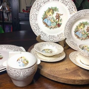 Può includere: Un set di stoviglie in porcellana bianca con bordi in filigrana dorata e illustrazioni centrali di figure in abiti d'epoca. Il set comprende una zuccheriera con coperchio, una ciotola su piattino, una tazza da tè su piattino e due piatti grandi.