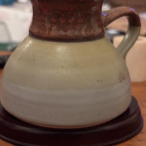 May include: A ceramic creamer pitcher with a rounded body and a single handle. The top section features a mottled reddish-brown glaze, while the lower portion transitions into a smooth, light beige finish with subtle horizontal ridges along the base.