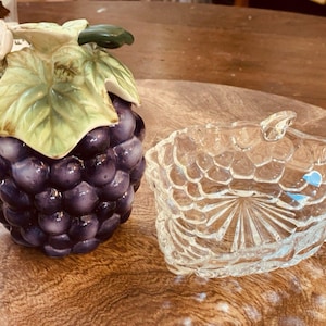 May include: A ceramic jar shaped like a cluster of purple grapes with green leaves and a small white flower on the lid, accompanied by a clear glass dish featuring a textured grape pattern on the exterior and a starburst design on the base.