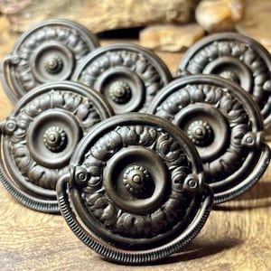 May include: A collection of circular metal drawer pulls featuring a dark bronze finish. Each pull has a central knob surrounded by a raised, textured ring with leaf-like patterns and a hinged, semi-circular handle attached to the outer edge.
