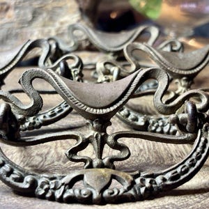 May include: A set of antique-style metal drawer pulls featuring a dark, weathered finish. Each pull has a curved, ornate backplate with intricate scrollwork and floral detailing, paired with a swinging, semi-circular handle loop.