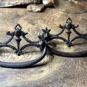 May include: Two dark metal drawer pulls featuring ornate, symmetrical backplates with intricate scrollwork and central finial details. Each pull has a curved, semi-circular handle attached to the decorative mounting plate, showcasing a weathered, aged finish.