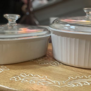 May include: Two white ceramic baking dishes with vertical ribbed sides and clear glass lids. One dish is oval-shaped and the other is round. Both lids feature a small, clear glass knob handle at the center for lifting the covers during cooking.