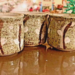 May include: Three ceramic cups with a speckled light green glaze and a hand-painted design of brown stems with white buds. The cups have a cylindrical shape with a slightly flared rim and a smooth, glossy finish on the exterior surface.