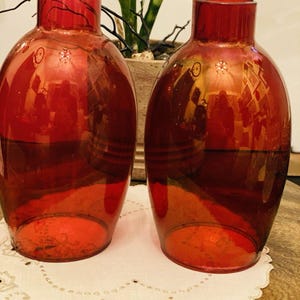 May include: Two matching translucent red glass vases with a rounded, bulbous body and a narrow, cylindrical neck. The glass has a smooth, polished finish that reflects light, creating a vibrant, deep crimson appearance across the surface.