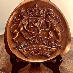 Op de afbeelding: Een driehoekige bronskleurige plaquette met een reliëf van het wapen van Luxemburg, inclusief twee leeuwen die een centraal schild geflankeerd door een kroon omringen. Onder het schild toont een banier de tekst GRAND DUCHE DE LUXEMBOURG.