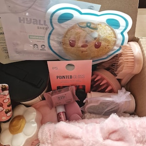 K&ouml;nnte beinhalten: Eine Box voller Sch&ouml;nheitsprodukte, darunter eine Gesichtsmaske, ein b&auml;renf&ouml;rmiger Schwamm, ein rosa Stirnband und eine Schachtel Pointed Gloss-N&auml;gel. Weitere Artikel sind Lipgloss, ein bl&uuml;tenf&ouml;rmiger Beh&auml;lter und eine B&uuml;rste.
