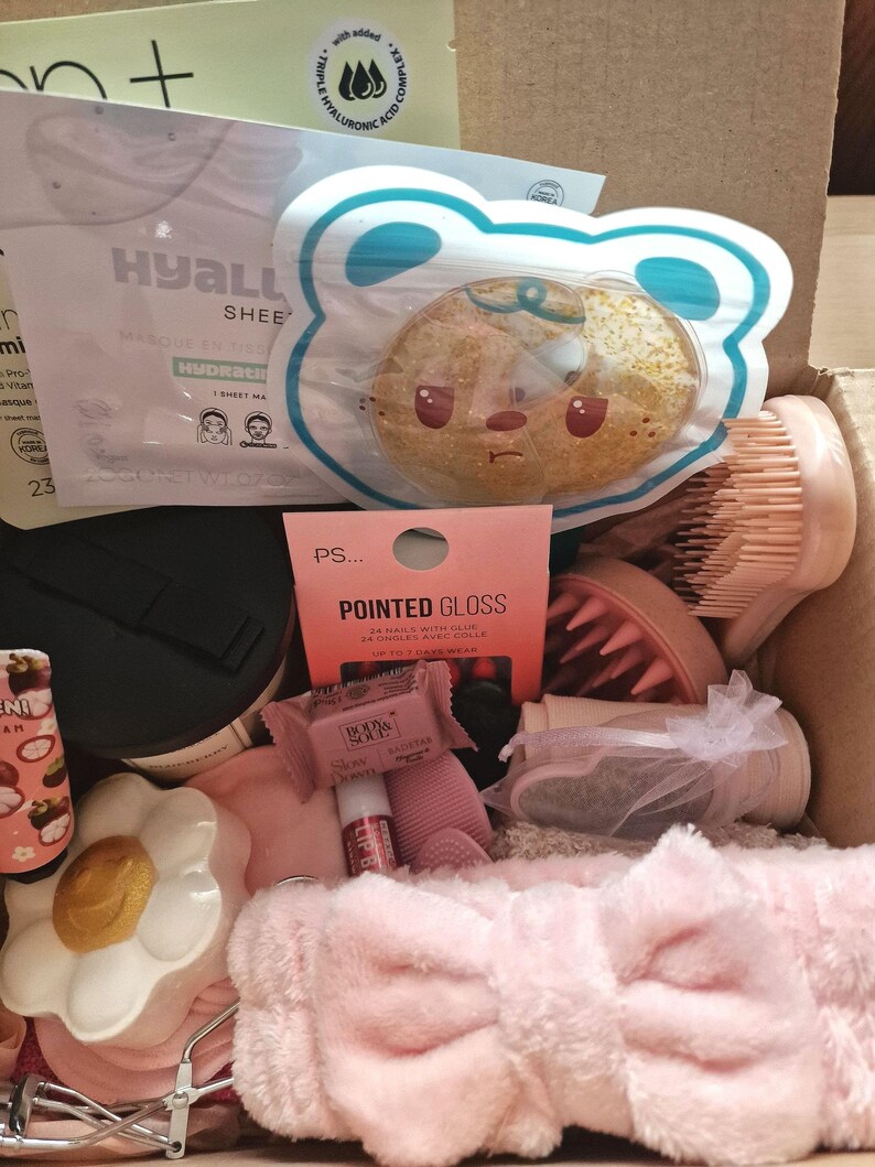 K&ouml;nnte beinhalten: Eine Box voller Sch&ouml;nheitsprodukte, darunter eine Gesichtsmaske, ein b&auml;renf&ouml;rmiger Schwamm, ein rosa Stirnband und eine Schachtel Pointed Gloss-N&auml;gel. Weitere Artikel sind Lipgloss, ein bl&uuml;tenf&ouml;rmiger Beh&auml;lter und eine B&uuml;rste.