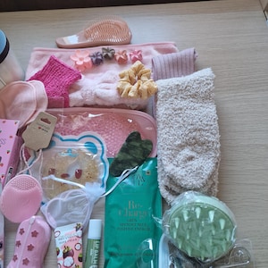 K&ouml;nnte beinhalten: Eine Sammlung von Pflegeprodukten in Rosa, Gr&uuml;n und Beige. Dazu geh&ouml;ren eine rosa Haarb&uuml;rste, Stirnb&auml;nder, Scrunchies, Socken, eine Gesichtsb&uuml;rste, ein rosa Stiefel, Lippenbalsam, ein gr&uuml;ner Gesichtsmassager und ein Paket mit der Aufschrift "Re-Charge".