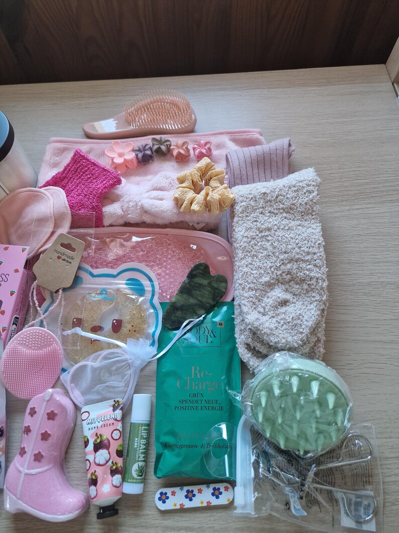 K&ouml;nnte beinhalten: Eine Sammlung von Pflegeprodukten in Rosa, Gr&uuml;n und Beige. Dazu geh&ouml;ren eine rosa Haarb&uuml;rste, Stirnb&auml;nder, Scrunchies, Socken, eine Gesichtsb&uuml;rste, ein rosa Stiefel, Lippenbalsam, ein gr&uuml;ner Gesichtsmassager und ein Paket mit der Aufschrift "Re-Charge".