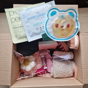 K&ouml;nnte beinhalten: Ein Karton voller Sch&ouml;nheitsprodukte, darunter Tuchmasken, eine Gesichtsb&uuml;rste, ein bl&uuml;tenf&ouml;rmiges Kompaktpuder und Wimpernzange. Die Box enth&auml;lt verschiedene Hautpflege- und Make-up-Artikel, die auf Selbstpflege ausgerichtet sind.