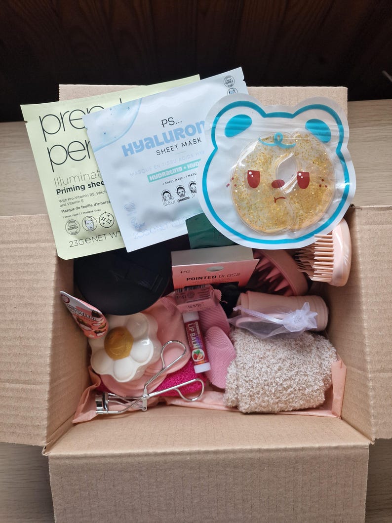 K&ouml;nnte beinhalten: Ein Karton voller Sch&ouml;nheitsprodukte, darunter Tuchmasken, eine Gesichtsb&uuml;rste, ein bl&uuml;tenf&ouml;rmiges Kompaktpuder und Wimpernzange. Die Box enth&auml;lt verschiedene Hautpflege- und Make-up-Artikel, die auf Selbstpflege ausgerichtet sind.