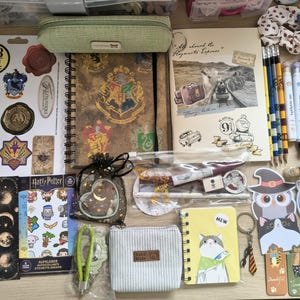 Może przedstawiać: Kolekcja artykułów papierniczych zawierająca notatnik spiralny Hogwart, arkusz naklejek Harry Potter, dziennik Hogwart Express, zielony piórnik, szary sztruksowy woreczek, różne karteczki samoprzylepne z kotami, ołówki, długopisy i zakładki.