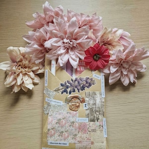 Może przedstawiać: Dekoracyjne opakowanie owinięte w brązowy papier, ozdobione różowymi i czerwonymi sztucznymi kwiatami. Opakowanie jest ozdobione papierem we wzory kwiatowe, naklejkami z tekstem i pieczęcią woskową. Tekst na naklejkach zawiera "Twilight whispers" i "Cherish the time."