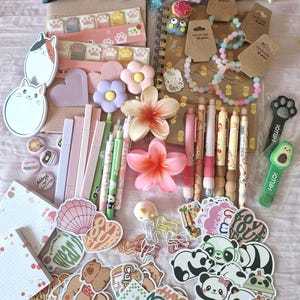 Może przedstawiać: Kolekcja artykułów papierniczych i akcesoriów w pastelowych kolorach. Zawiera naklejki, długopisy, bransoletki, gumki do włosów i ozdobne kwiaty. Przedmioty są ułożone na jasnoróżowej powierzchni, tworząc atrakcyjny wizualnie asortyment.