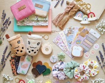 May include: A collection of stationery and accessories including two plush boba tea pouches, a donut charm, heart-shaped leather pieces, fabric scrunchies with cherry and floral prints, macrame rainbow keychains, and various stickers and bookmarks.