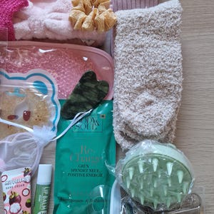 K&ouml;nnte beinhalten: Eine Sammlung von Selbstpflegeartikeln, darunter eine rosa Augenmaske, ein Jade-Gua-Sha-Werkzeug, Handcreme, Lippenbalsam und ein gr&uuml;ner Kopfhautmassageger&auml;t. Weitere Artikel sind ein flauschiges Stirnband, Scrunchies, Socken und eine Packung mit dem Text "Re-Charge".