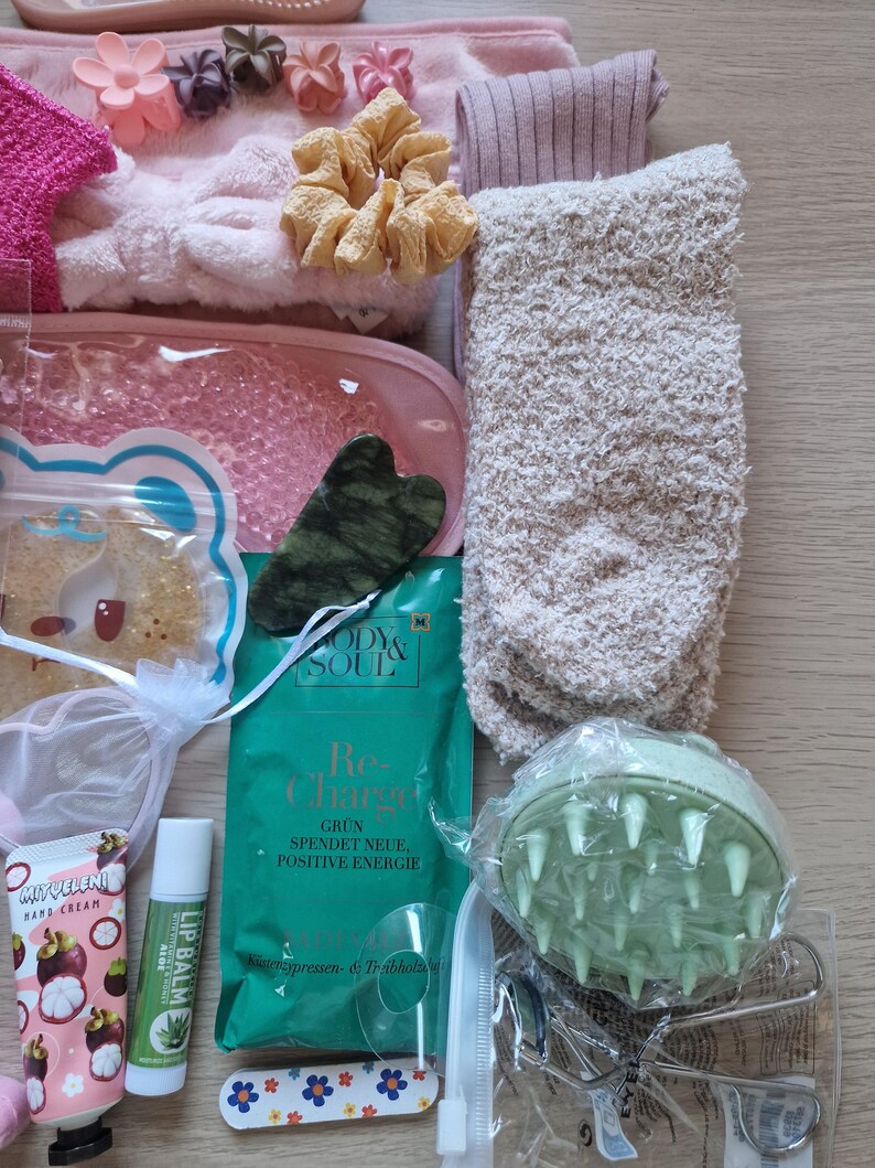 K&ouml;nnte beinhalten: Eine Sammlung von Selbstpflegeartikeln, darunter eine rosa Augenmaske, ein Jade-Gua-Sha-Werkzeug, Handcreme, Lippenbalsam und ein gr&uuml;ner Kopfhautmassageger&auml;t. Weitere Artikel sind ein flauschiges Stirnband, Scrunchies, Socken und eine Packung mit dem Text "Re-Charge".