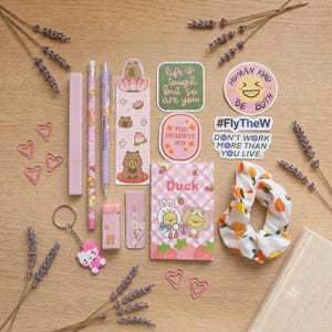 Può includere: Un set di articoli di cancelleria che include un astuccio viola chiaro, due matite, un segnalibro con illustrazioni di capibara, un piccolo taccuino con grafiche di anatre e orsi, una gomma rosa, un portachiavi, adesivi e un elastico per capelli bianco con stampa di arance.