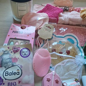 K&ouml;nnte beinhalten: Eine Sammlung von Beauty- und Pflegeprodukten, darunter eine Duftkerze, Gesichtsmasken, ein Nagelset und eine rosa Gesichtsreinigungsb&uuml;rste. Weitere Artikel sind ein rosa Stirnband, Haarschmuck und eine Handcreme. Die Produkte sind auf einer Holzoberfl&auml;che angeordnet.