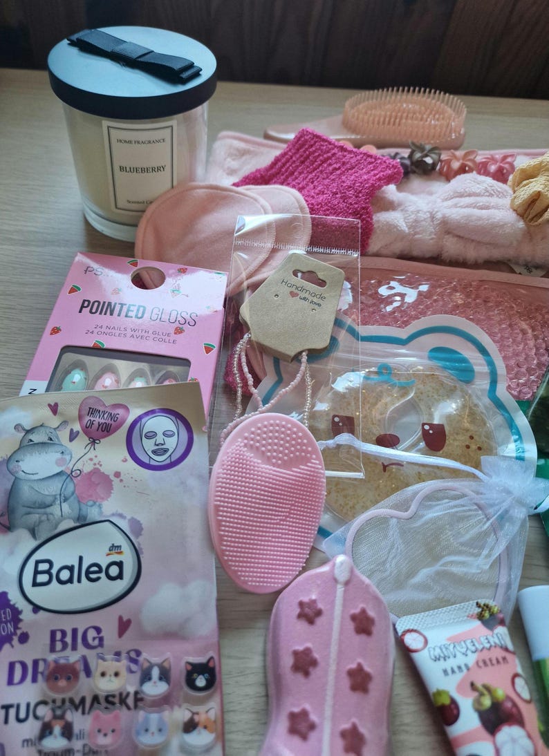 K&ouml;nnte beinhalten: Eine Sammlung von Beauty- und Pflegeprodukten, darunter eine Duftkerze, Gesichtsmasken, ein Nagelset und eine rosa Gesichtsreinigungsb&uuml;rste. Weitere Artikel sind ein rosa Stirnband, Haarschmuck und eine Handcreme. Die Produkte sind auf einer Holzoberfl&auml;che angeordnet.