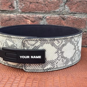 以下が含まれることがあります： グレーと白のヘビ柄のウェイトリフティングベルト。ベルトには、白い文字で「YOUR NAME」と書かれた黒いバックルが付いています。ベルトの内側はネイビーブルーです。