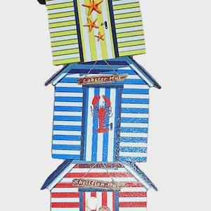 May include: A vertical decorative stack of three beach huts featuring yellow, blue, and red stripes. The huts are labelled Starfish Hut, Lobster Hut, and Shellfish Hut. The base includes two miniature beach chairs, a red bucket, and a beach ball.